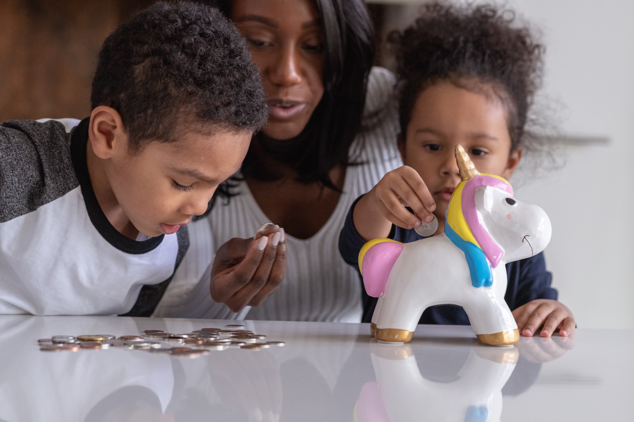 Woman overlooking two small kids at a table. The kids are looking at coins on the table and putting them into a unicorn piggy bank.
